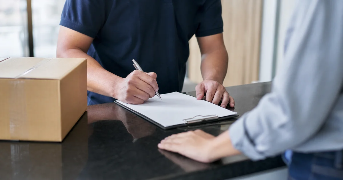 Persona firmando una nota de entrega en una planilla sobre un mesón de recepción, sin mostrar el rostro